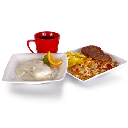Two white plates with breakfast scrambled eggs hash browns and a sausage patty on one plate plus a creamy chicken dish with gravy and an orange wedge on the other a red mug sits behind