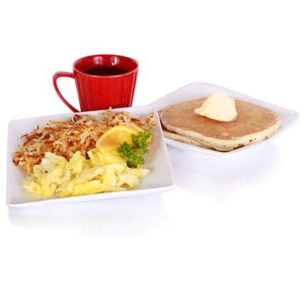 Two white plates of breakfast scrambled eggs hash browns bacon with an orange slice on the first plate a buttered pancake on the second plate and a red mug of coffee in the background