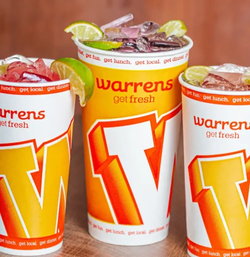 Three Warrens beverage cups with big orange 'W' logo and lime wedges on a wooden table, filled with ice.