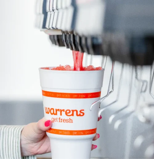 Hand holding a white cup labeled 'Warrens get fresh' as red slush pours from a drink machine into it