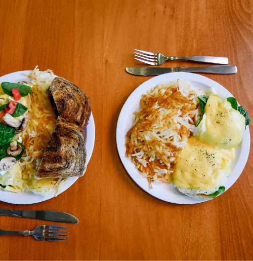 Two plates of breakfast on a wooden table: left plate has a veggie scramble with spinach, mushrooms, tomatoes and a crusty loaf on top of hash browns; right plate features eggs Benedict with hollandaise over greens and hash browns.