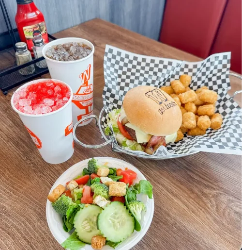 Hamburger with cheese and lettuce in a basket, with crispy tater tots, a cucumber-broccoli salad, and two drinks on a wooden table.