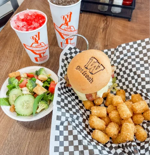 Burger with cheese on a bun, a side of crispy tater tots in checkered liner, and a small salad on the side; two cups with ice drinks in the background on a wooden table.